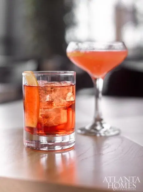 Two red cocktails with ice and garnishes on a wooden table, with the Atlanta Homes & Lifestyles logo in the lower right corner.