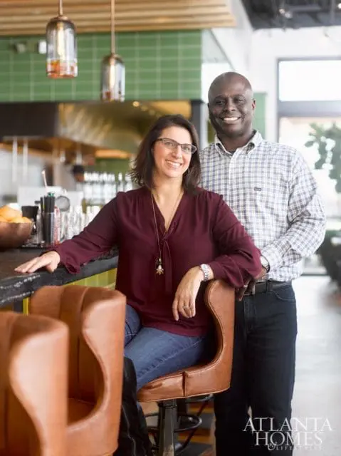 A smiling woman wearing glasses and a maroon blouse sits on a brown leather bar stool, while a smiling man in a plaid shirt stands behind her.