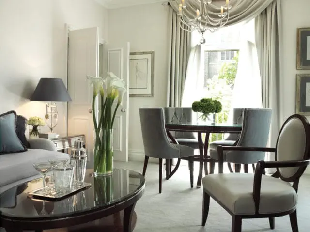 Living and dining area with gray upholstered chairs, a dark wood dining table, a crystal chandelier, and a vase of white calla lilies on a coffee table.
