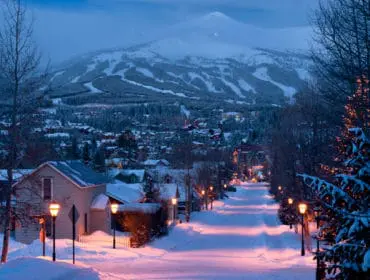 Snow-covered street in a mountain town at dusk, with houses, streetlights, bare trees, and a ski slope on a distant mountain