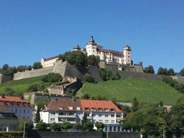 Marienberg Fortress in Wurzburg, Germany, a large white castle with red-tiled roofs and multiple towers, perched atop a steep, green, vineyard-covered hill with stone fortifications.