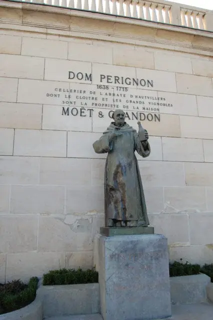 Statue of Dom Perignon holding a bottle in front of a stone wall inscribed with text about him and Moët & Chandon.
