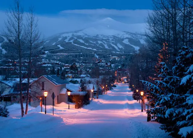Snowy street lined with houses and lampposts leading to a mountain village at dusk, with ski slopes visible on the mountain