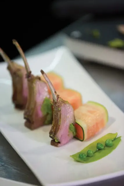Three racks of lamb with green herb paste, served with sliced cantaloupe and a pea pod with four peas on a white rectangular plate.