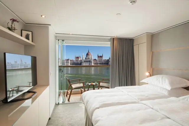 Bedroom with two beds, a TV, and a balcony overlooking a river and a grand building.