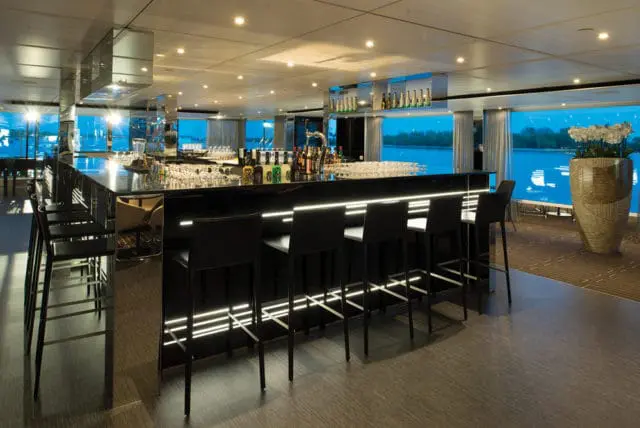 Modern bar with black counter, illuminated by white LED strips, and black bar stools. Large windows overlook a body of water at dusk.