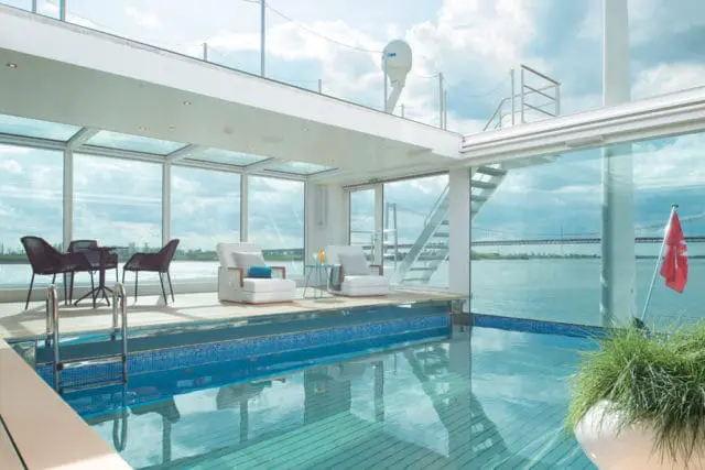 Indoor pool with blue tiled accents, white lounge chairs, and a glass-enclosed ceiling overlooking a river and bridge.