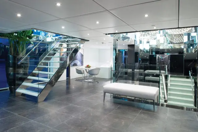 Modern interior with a glass and chrome staircase, gray tiled floor, white bench, and mirrored walls. A small dining set is visible in the background.