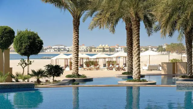 Resort pool with palm trees, beach cabanas, and buildings in the background under a clear blue sky.