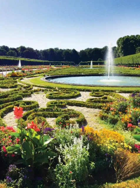 Formal garden with a circular fountain, manicured hedges, and colorful flower beds under a clear blue sky.