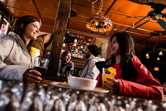 Two women and two men sitting at a bar, smiling and holding drinks. The bar has a rustic wooden interior with a decorative stained-glass lamp hanging from the ceiling.