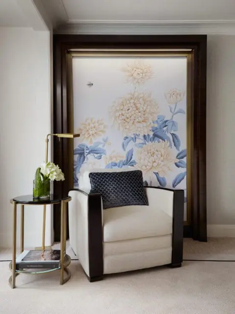 Cream upholstered armchair with dark wood trim and a dark patterned pillow, next to a small round side table with a glass vase of white flowers and a book. Behind the chair is a large framed artwork of pale yellow chrysanthemums and blue foliage.