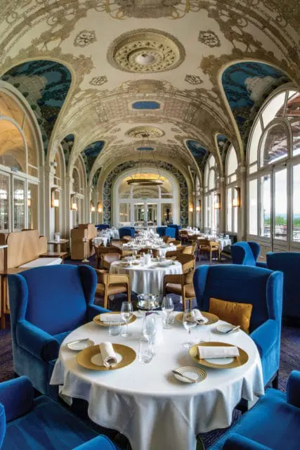 Elegant dining room with ornate vaulted ceilings, blue velvet armchairs, and white tablecloths set with gold chargers.