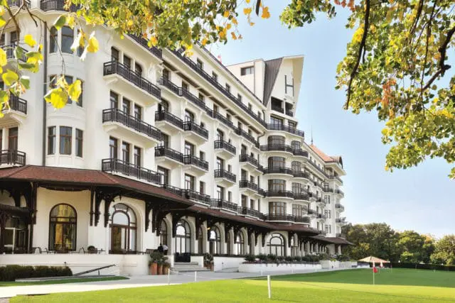 Grand white hotel with multiple balconies and arched windows, set against a clear blue sky with autumn leaves in the foreground and a manicured lawn with golf flags in the background.