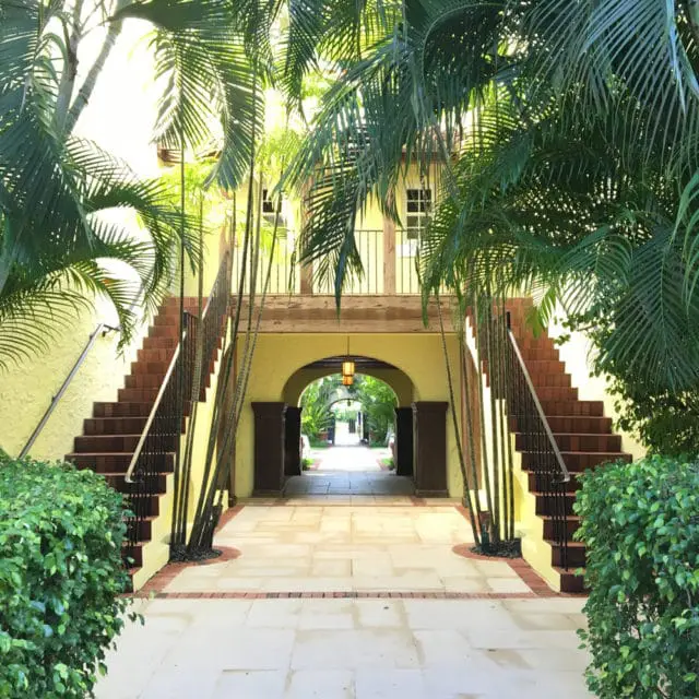 Yellow stucco building with arched entryway, flanked by brick stairs and palm trees. A walkway with large stone pavers leads through the archway.
