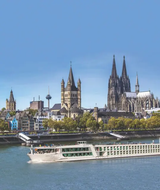 A white cruise ship sails on a blue river in front of the Cologne skyline, featuring several historic churches and buildings under a clear sky.