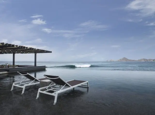 Infinity pool overlooking the ocean with a wave breaking in the distance. Two lounge chairs sit by the pool, and a covered bar area is to the left.