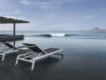 Infinity pool overlooking the ocean with a wave breaking in the distance. Two lounge chairs sit by the pool, with a covered bar area to the left.
