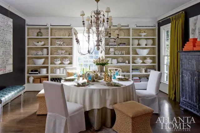 Dining room with a round table set for four, slipcovered chairs, a large ornate chandelier, and floor-to-ceiling bookshelves with decorative items.