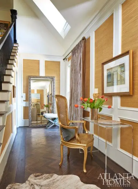 Foyer with grasscloth wallpaper, a gold cane chair, a large ornate mirror, and a clear console table with tulips.