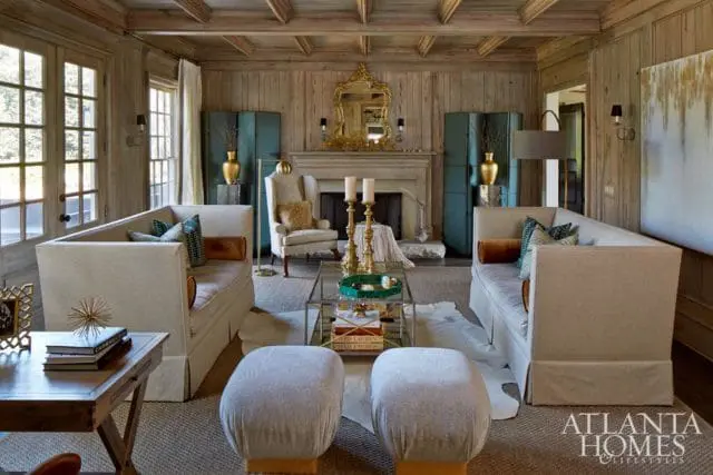 Living room with wood-paneled walls and beamed ceiling, featuring two cream slipcovered sofas, a fireplace with a gold ornate mirror, and two upholstered ottomans.