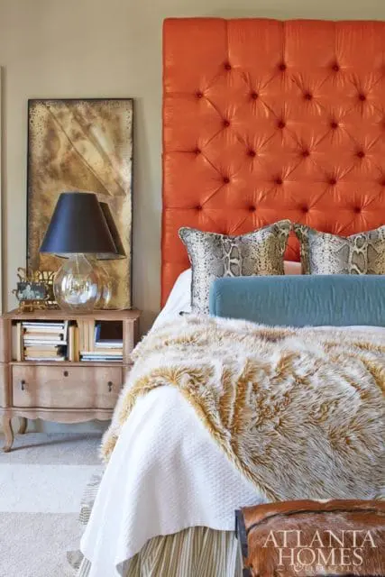 Master bedroom with a tufted orange headboard, snake print pillows, a teal bolster pillow, and a faux fur throw blanket. A wooden nightstand holds a lamp and books.