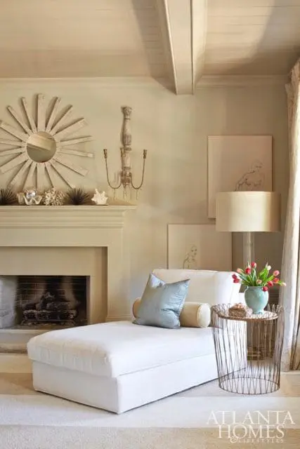 Master bedroom with a white chaise lounge, light gray walls, a cream fireplace with a sunburst mirror above, and a side table with a vase of tulips.