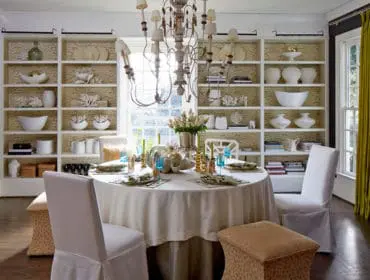 Dining room with a round table set for a meal, white slipcovered chairs, and floor-to-ceiling bookshelves filled with decorative objects.