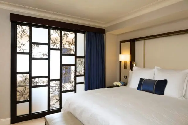 Junior suite bedroom with a king-size bed, white linens, and a dark blue accent pillow. A decorative glass partition with floral motifs separates the room from a blue curtained area.