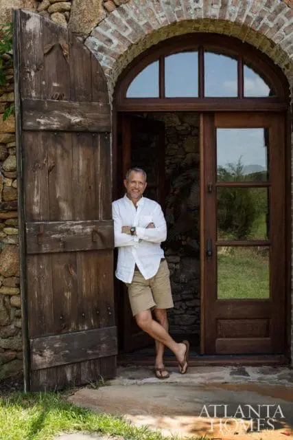Man in white shirt and khaki shorts leaning against a rustic wooden door with stone wall and arched brick entryway.