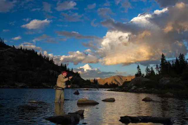Man fly fishing in a lake with mountains and pine trees in the background under a dramatic cloudy sky at sunset.