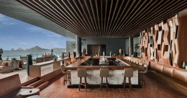 Modern bar with a large wooden counter, leather seating, and a slatted wood ceiling, overlooking the ocean and distant islands.