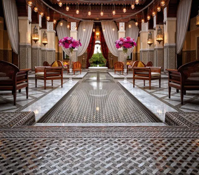 Ornate courtyard interior with a long water feature, intricate tile flooring, wooden furniture, and decorative lanterns hanging from the ceiling.