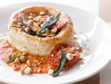 Round pastry dish with layered eggplant, tomato, and herbs, topped with chopped nuts and served with a red sauce on a white plate.