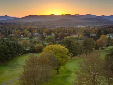 Golf course with rolling green fairways and trees in autumn foliage, with mountains and a sunset in the background.