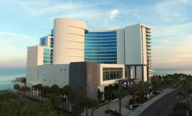 Modern white hotel with curved glass facade and balconies overlooking the ocean, lined with palm trees and a coastal road.