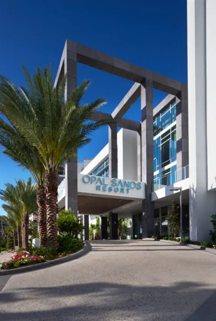 Opal Sands Resort entrance with modern architecture, geometric concrete overhang, palm trees, and a bright blue sky.