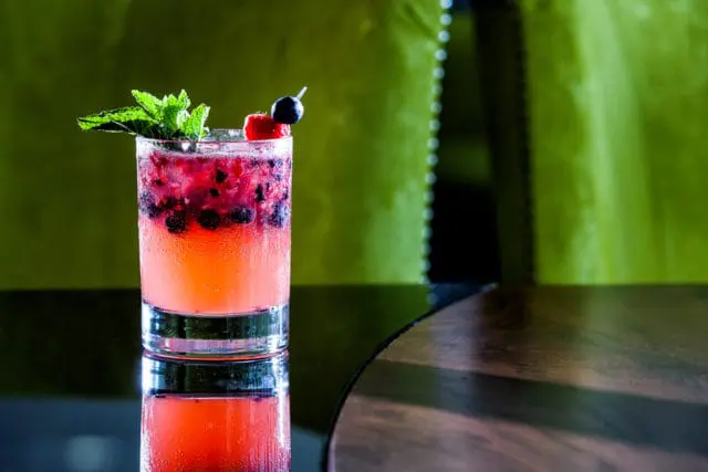 A tall glass of pink cocktail with blueberries and raspberries, garnished with mint, a strawberry slice, and a blueberry.