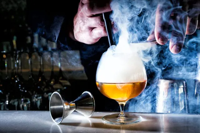Bartender smoking a cocktail in a snifter glass with a jigger and shaker in the background.