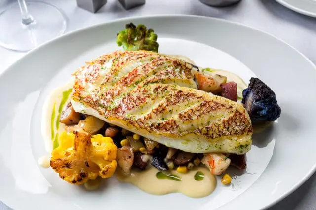 Pan-seared hogfish fillet with red skin, served over roasted vegetables including corn, potatoes, and cauliflower, with a creamy sauce and herb garnish.