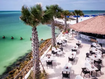 Beachside outdoor bar with thatched roof, white bar stools, and numerous tables and chairs on a sandy patio overlooking the turquoise ocean and palm trees.