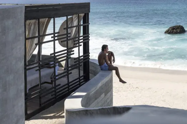 Man sitting on a concrete wall overlooking a beach and ocean, next to an open-air cabana with sheer curtains.