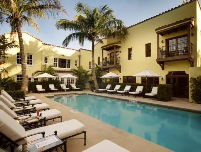 Yellow stucco building with Spanish tile roof and dark wood balconies surrounding a bright blue swimming pool with lounge chairs and umbrellas
