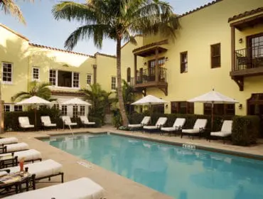 Yellow stucco building with Spanish tile roof surrounding a bright blue swimming pool with white lounge chairs and umbrellas.