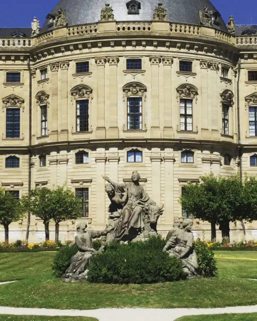 Baroque palace with ornate facade and dome, featuring a central sculpture group of figures and a bull in a manicured garden setting.