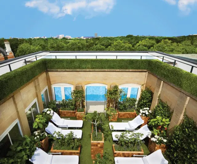 Rooftop garden with white lounge chairs, potted plants, and manicured hedges overlooking a cityscape and trees.