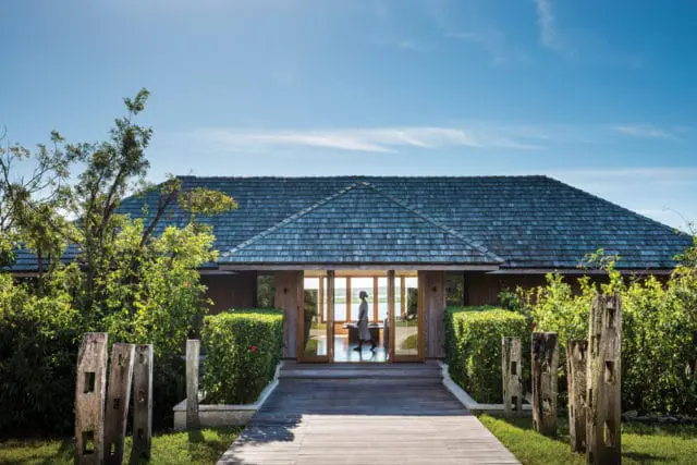 Wooden walkway leading to a single-story building with a dark wood shingle roof and large glass doors. Lush green bushes flank the entrance, and weathered wooden posts stand in the foreground.