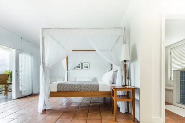 Bedroom with a wooden four-poster bed draped in white sheer fabric, terracotta tile flooring, and a wooden side table with a white lamp.