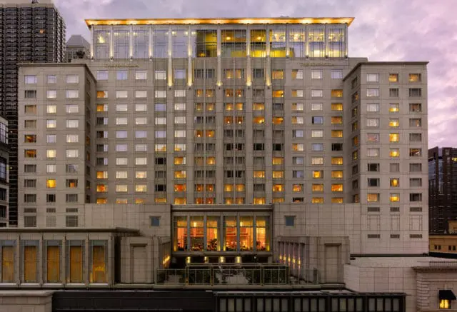 The Peninsula hotel at dusk, a tall building with many windows lit from within, featuring a glass-enclosed top section and ornate architectural details.