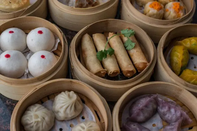 Assortment of dim sum in bamboo steamers: white buns with red dots, spring rolls, yellow dumplings, purple dumplings, and soup dumplings.